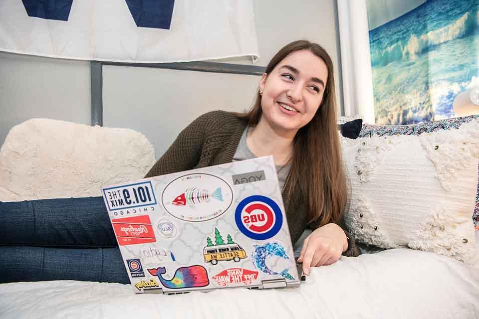 A resident reclines on her side on a bed with pillows, she's smiling at something off to the side. A laptop with many stickers is in front of her.